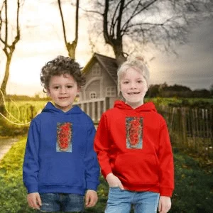 Due bambini sorridenti posano all'aperto davanti a una piccola casa di legno in un paesaggio rurale al tramonto. Il bambino a sinistra indossa una felpa con cappuccio blu reale, mentre la bambina a destra indossa una felpa con cappuccio rossa. Entrambe le felpe presentano una stampa artistica rettangolare al centro con toni caldi e motivi floreali o astratti.