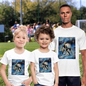 Un uomo e due bambini  posano insieme su un campo sportivo all'aperto. Tutti e tre indossano magliette bianche a maniche corte con la stessa stampa artistica quadrata al centro, che raffigura una scena dinamica in tonalità di blu e ocra. Lo sfondo mostra un prato verde e altre persone sfocate durante un'attività sportiva.