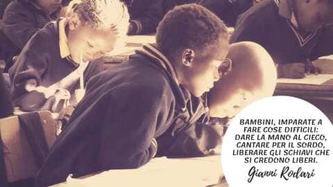 Fotografia d'epoca in bianco e nero virata seppia che ritrae un gruppo di bambini in un'aula scolastica, concentrati mentre scrivono. In basso a destra, un cerchio bianco contiene la citazione di Gianni Rodari: "Bambini, imparate a fare cose difficili: dare la mano al cieco, cantare per il sordo, liberare gli schiavi che si credono liberi".